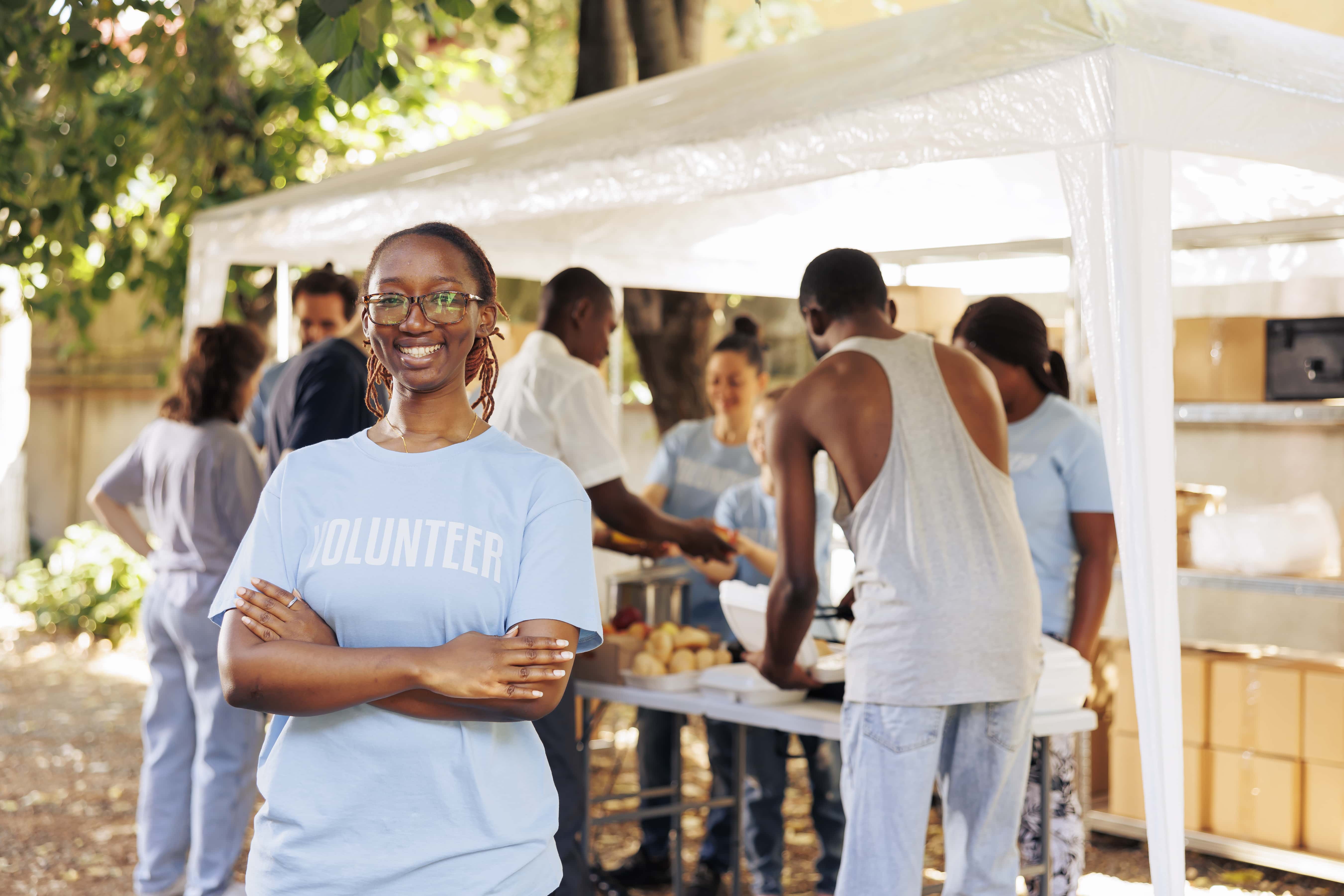 young black woman with glasses stands outdoors arms crossed looking camera diverse group volunteers supports non profit program dedicated hunger relief helping needy